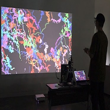 A visitor stands in a dark gallery beside a microscope setup, facing a large projection filled with tangled, multicolored trails.